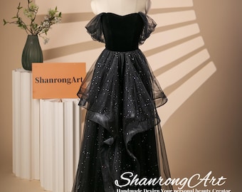 May include: A black off-the-shoulder gown with a velvet bodice and a tulle skirt with glitter. The skirt is layered and has a ruffled design. The gown is on a mannequin against a light brown background.