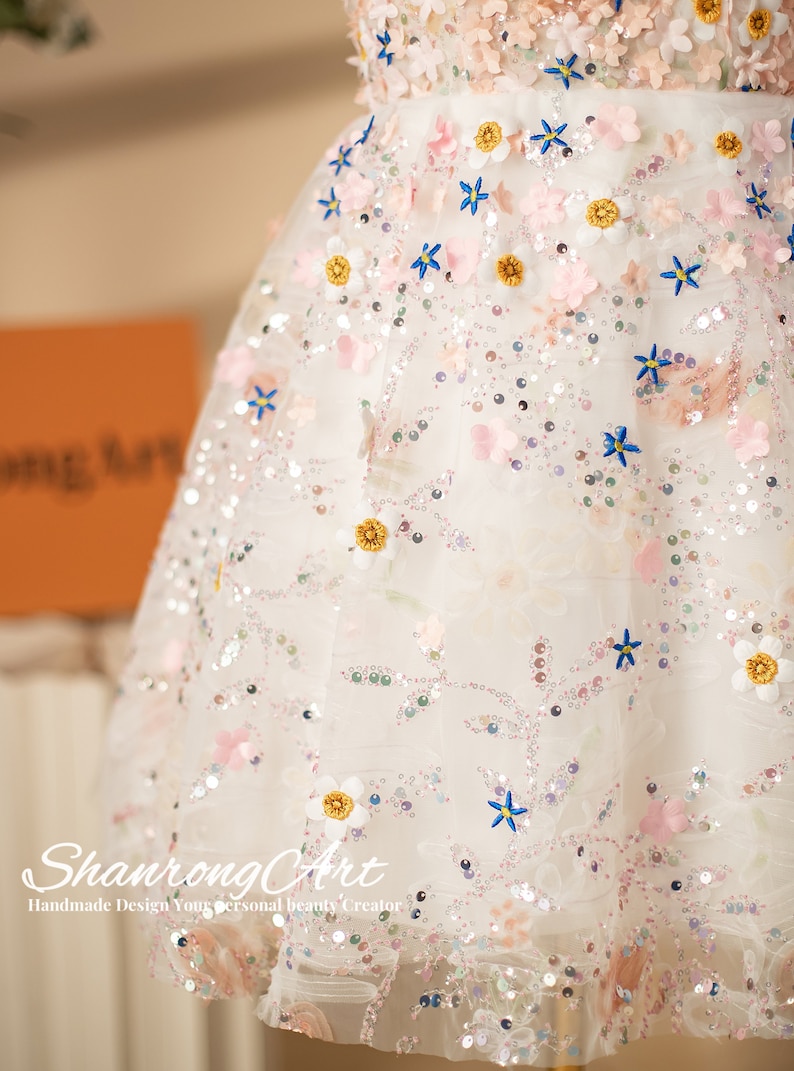 K&ouml;nnte beinhalten: Ein wei&szlig;es T&uuml;llkleid mit rosa und blauen Blumenverzierungen. Das Kleid ist mit Pailletten besetzt und hat einen vollen Rock.