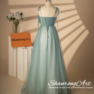 May include: A light blue tulle dress with a lace-up back and thin straps. The dress has a flowing skirt and is photographed on a mannequin against a light brown background.