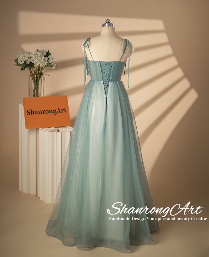 May include: A light blue tulle dress with a lace-up back and thin straps. The dress has a flowing skirt and is photographed on a mannequin against a light brown background.