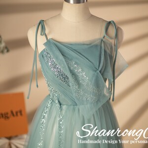 May include: A light blue tulle dress with silver sequins. The dress has a square neckline and thin straps that tie at the shoulders. The dress is on a white mannequin.