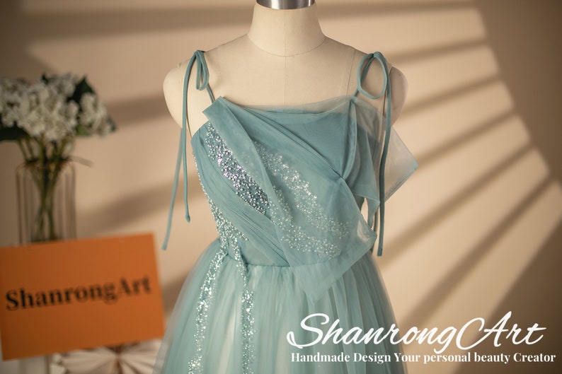 May include: A light blue tulle dress with silver sequins. The dress has a square neckline and thin straps that tie at the shoulders. The dress is on a white mannequin.