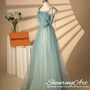 May include: A light blue tulle dress with a square neckline and thin straps. The dress has a fitted bodice and a flowing skirt. The dress is decorated with small sequins. The dress is on a mannequin and the background is a light beige color. The text "ShanrongArt Handmade Design Your personal beauty Creator" is at the bottom of the image.