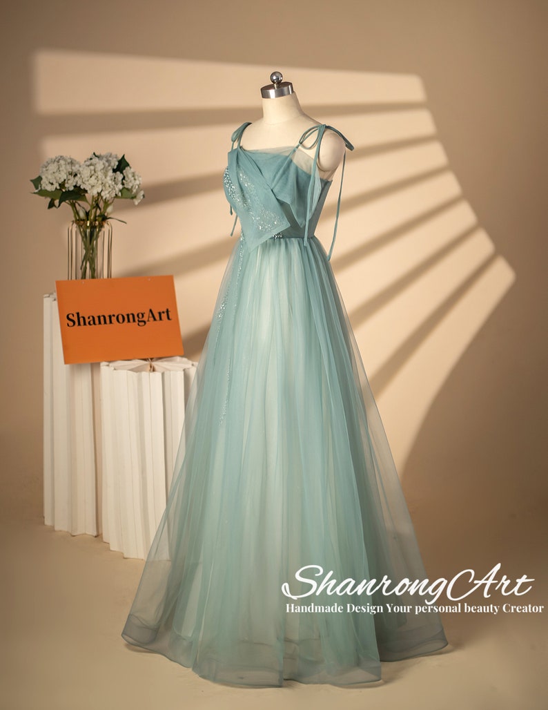 May include: A light blue tulle dress with a square neckline and thin straps. The dress has a fitted bodice and a flowing skirt. The dress is decorated with small sequins. The dress is on a mannequin and the background is a light beige color. The text "ShanrongArt Handmade Design Your personal beauty Creator" is at the bottom of the image.