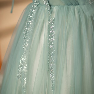 May include: A light blue tulle dress with silver embellishments. The dress has a fitted bodice and a flowing skirt. The skirt is decorated with silver sequins and beads. The dress is perfect for a special occasion.