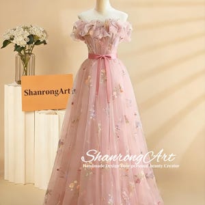 Könnte beinhalten: Ein hellrosa schulterfreies Kleid mit floralen Stickereien und einem passenden Schleifengürtel. Das Kleid hat einen fließenden Tüllrock und wird auf einer Schaufensterpuppe präsentiert. Der Text "ShanrongArt" ist sichtbar.