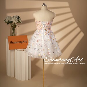 K&ouml;nnte beinhalten: Ein wei&szlig;es tr&auml;gerloses Kleid mit Blumenmuster und einem T&uuml;llrock. Das Kleid befindet sich auf einer Puppe und hat einen Rei&szlig;verschluss im R&uuml;cken. Das Kleid ist mit kleinen Blumen und Pailletten verziert.