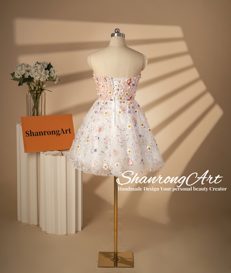 K&ouml;nnte beinhalten: Ein wei&szlig;es tr&auml;gerloses Kleid mit Blumenmuster und einem T&uuml;llrock. Das Kleid befindet sich auf einer Puppe und hat einen Rei&szlig;verschluss im R&uuml;cken. Das Kleid ist mit kleinen Blumen und Pailletten verziert.