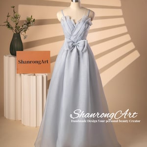 May include: A light blue sleeveless gown with a sweetheart neckline and a large bow at the waist. The gown is made of a sheer fabric and has a flowing skirt. The gown is on a mannequin against a light brown background.