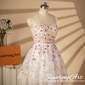 K&ouml;nnte beinhalten: Ein wei&szlig;es T&uuml;llkleid mit einem herzf&ouml;rmigen Ausschnitt und einem Blumendesign. Das Kleid ist mit rosa, blauen und wei&szlig;en Blumen und Pailletten bedeckt. Das Kleid befindet sich auf einer Puppe und hat eine kurze, A-f&ouml;rmige Silhouette.