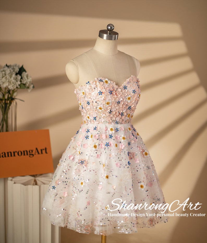 K&ouml;nnte beinhalten: Ein wei&szlig;es T&uuml;llkleid mit einem herzf&ouml;rmigen Ausschnitt und einem Blumendesign. Das Kleid ist mit rosa, blauen und wei&szlig;en Blumen und Pailletten bedeckt. Das Kleid befindet sich auf einer Puppe und hat eine kurze, A-f&ouml;rmige Silhouette.