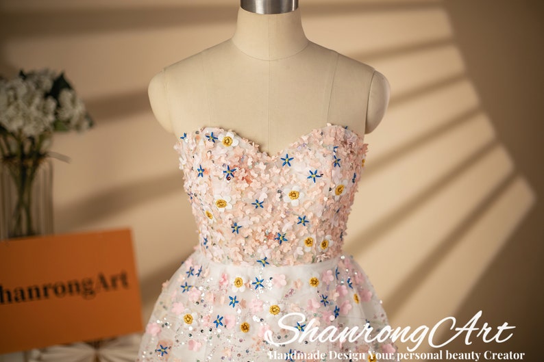K&ouml;nnte beinhalten: Ein wei&szlig;es T&uuml;llkleid ohne Tr&auml;ger mit einem rosa-wei&szlig;en Blumendesign. Das Kleid ist mit Pailletten und kleinen Blumen verziert. Das Kleid befindet sich auf einer wei&szlig;en Puppe.