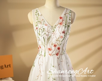 Puede incluir: Un vestido blanco sin mangas con escote en V y bordado floral. El vestido tiene una tela de tul y una falda fluida. El bordado presenta flores rosas, verdes y blancas.