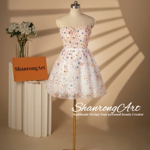 K&ouml;nnte beinhalten: Ein wei&szlig;es tr&auml;gerloses Kleid mit einem rosa-wei&szlig;en Blumendesign. Das Kleid ist aus T&uuml;ll und hat eine kurze, A-f&ouml;rmige Silhouette. Das Kleid ist mit Pailletten und Perlen verziert.