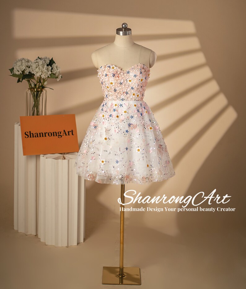 K&ouml;nnte beinhalten: Ein wei&szlig;es tr&auml;gerloses Kleid mit einem rosa-wei&szlig;en Blumendesign. Das Kleid ist aus T&uuml;ll und hat eine kurze, A-f&ouml;rmige Silhouette. Das Kleid ist mit Pailletten und Perlen verziert.