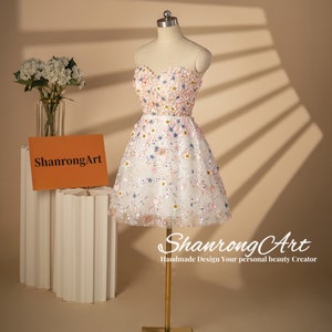 K&ouml;nnte beinhalten: Ein wei&szlig;es, tr&auml;gerloses Minikleid mit rosa und blauen Blumenstickereien und Pailletten. Das Kleid befindet sich auf einer Puppe und hat einen herzf&ouml;rmigen Ausschnitt.