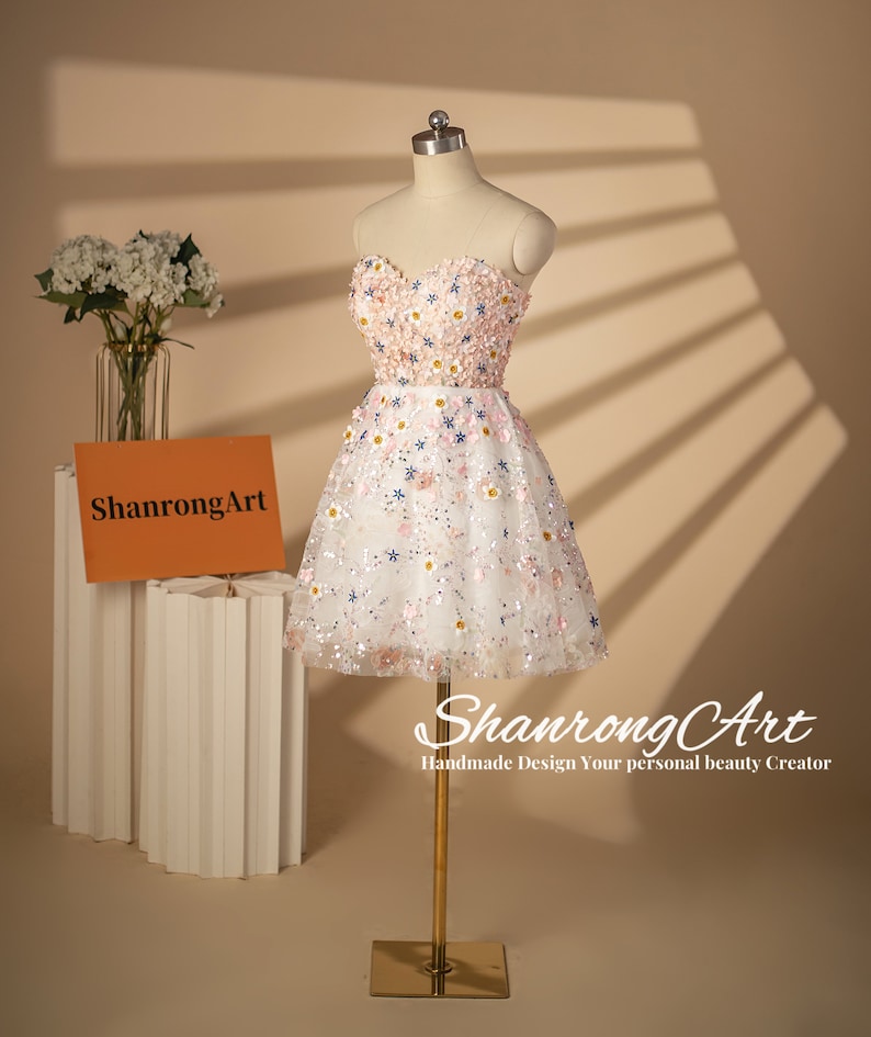 K&ouml;nnte beinhalten: Ein wei&szlig;es, tr&auml;gerloses Minikleid mit rosa und blauen Blumenstickereien und Pailletten. Das Kleid befindet sich auf einer Puppe und hat einen herzf&ouml;rmigen Ausschnitt.