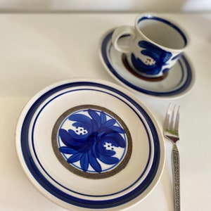 Könnte beinhalten: Ein weißer Keramikteller mit einem blauen und goldenen Blumendesign. Der Teller hat einen blauen Rand und ist von einer weißen Untertasse und einer blauen und weißen Teetasse umgeben.