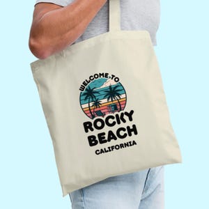 May include: Cream tote bag with a circular graphic showing palm trees, a beach scene, and the text "WELCOME TO ROCKY BEACH CALIFORNIA". The bag is held by a person.