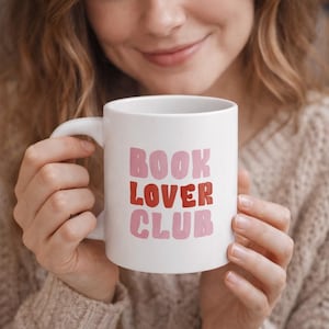 Könnte beinhalten: Weiße Keramik-Tasse mit dem Text "BOOK LOVER CLUB" in rosa und roter Schrift. Die Tasse hat einen gebogenen Henkel und wird von einer Person gehalten. Ein tolles Geschenk für Leseratten.