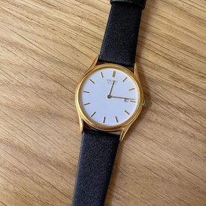 Puede incluir: Un reloj Seiko Quartz dorado con esfera blanca y correa de cuero negra.
