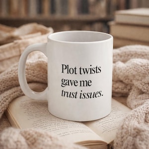 May include: White ceramic mug with the text "Plot twists gave me trust issues" in black. The mug is on top of an open book and a beige knitted blanket. The background is blurred, suggesting a cozy setting.