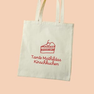Könnte beinhalten: Eine weiße Stofftasche mit rotem Text und einer roten Zeichnung eines Kirschkuchens. Der Text lautet "Tante Mathildas Kirschkuchen".