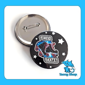May include: A black button with the text "TIME 2 SKATE!" and an illustration of roller skates. The button has a silver pin back and is surrounded by white stars. The Tacey Shop logo is in the bottom right corner.