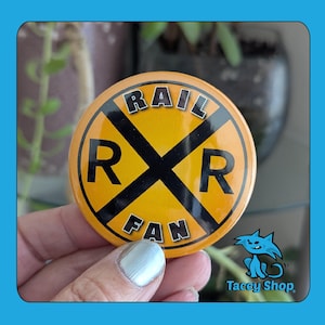 May include: A round, yellow button with a black railroad crossing symbol and the words "RAIL FAN" in white. The button is held by a person with a silver painted fingernail. The button has a blue border and a logo for "Tacey Shop".