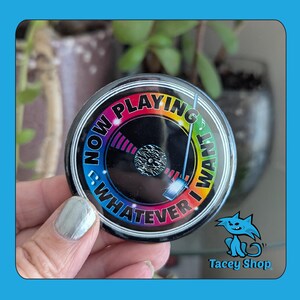 May include: A round, black and rainbow-colored button with the text "NOW PLAYING WHATEVER I WANT". The button features a vinyl record design with a needle and a rainbow gradient around the edge.