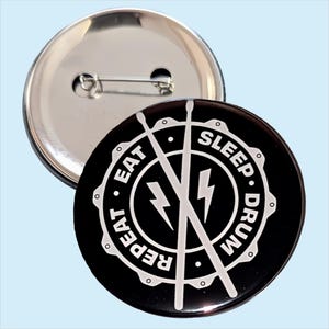 May include: A round pin-back button featuring a black background with a white graphic of two crossed drumsticks over a drum outline. The text EAT SLEEP DRUM REPEAT is printed in white around the circular border, with two lightning bolt icons in the center.