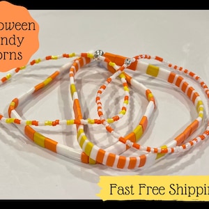 Può includere: Tre braccialetti colorati realizzati con perline bianche, arancioni e gialle. I braccialetti sono disposti in cerchio su una superficie bianca. Il testo "Halloween Candy Corns" è visibile nell'angolo in alto a sinistra dell'immagine. Il testo "Fast Free Shipping" è visibile nell'angolo in basso a destra dell'immagine.