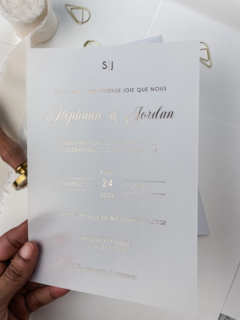 May include: A white wedding invitation with gold foil lettering. The invitation reads "C'est avec une immense joie que nous Stephanie & Jordan Vous invitons joyeusement &agrave; la c&eacute;l&eacute;bration de notre mariage le Samedi 24 Aout 2024 15h30 &agrave; l'h&ocirc;tel de ville de Bretigny-sur-Orge Merci de r&eacute;pondre au 06 00 00 00 Reception apr&egrave;s la c&eacute;r&eacute;monie."