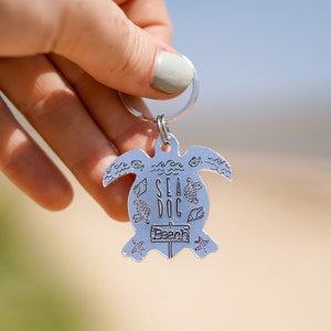 May include: Silver turtle keyring with the words "Sea Dog Beach" engraved on it. The turtle has a small sign that says "Beach" and is surrounded by small sea life designs.