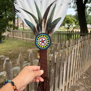 May include: A white and green feather fan with a beaded handle. The handle is brown leather with a beaded circle at the top. The circle has a blue, yellow, red, and black design.
