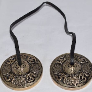 May include: A pair of small, round, brass cymbals with intricate designs and the word "Om" inscribed on them. The cymbals are connected by a black cord.