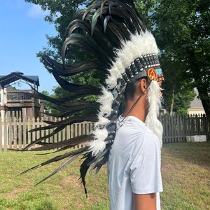 May include: A person wearing a black and white feathered headdress with a colorful beaded band. The headdress has a white fur trim and is worn over a white t-shirt.