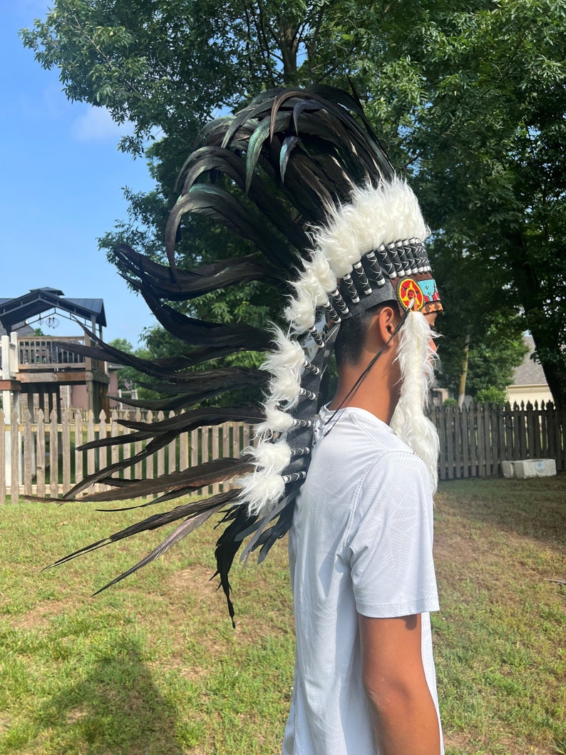 May include: A person wearing a black and white feathered headdress with a colorful beaded band. The headdress has a white fur trim and is worn over a white t-shirt.