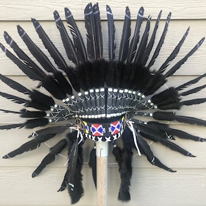 May include: A black feather headdress with a black and white beaded band and a red, white, and blue beaded band. The headdress is mounted on a wooden stick.