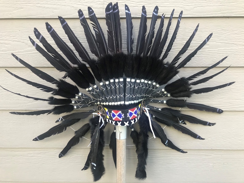 May include: A black feather headdress with a black and white beaded band and a red, white, and blue beaded band. The headdress is mounted on a wooden stick.