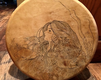 May include: A round, light brown leather drum with a black and white design of a woman with long hair, fish, and flowers. The drum is on a wooden floor.