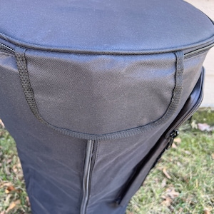 May include: Black cylindrical bag with a zippered top and side pocket. The bag is made of a durable, textured fabric and sits on a grassy surface. The top of the bag is round, and the sides are straight.