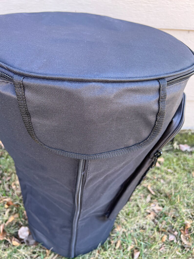 May include: Black cylindrical bag with a zippered top and side pocket. The bag is made of a durable, textured fabric and sits on a grassy surface. The top of the bag is round, and the sides are straight.