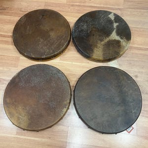 May include: Four round, hand drums with natural hide drumheads. The drumheads are various shades of brown and tan, with visible textures and patterns. The drums are arranged in a square formation.