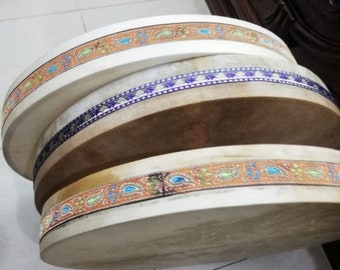 May include: A set of three round, white, hand drums with decorative, colorful, paisley-patterned trim. The drums are stacked on top of each other.