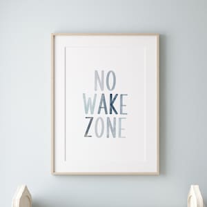May include: A white crib with a white and gray quilt, a rocking horse, and a wicker chair with a white cushion. A framed print with the text "NO WAKE ZONE" hangs on the wall above the crib.