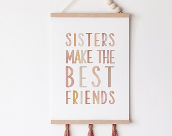 May include: A framed print with the text "SISTERS MAKE THE BEST FRIENDS" in various earth-tone colors. The print is suspended by a wooden rod and a string with wooden beads, and has tassels at the bottom.