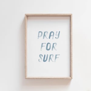 Puede incluir: Una impresión enmarcada con el texto "Pray for Surf" en letras azules sobre un fondo blanco. El marco es de madera clara.