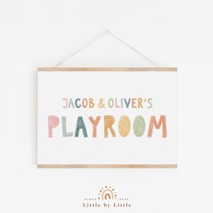 May include: A framed print with the text "Jacob & Oliver's Playroom" in colorful, hand-drawn letters. The print has a light wood frame and is hanging on a white wall.