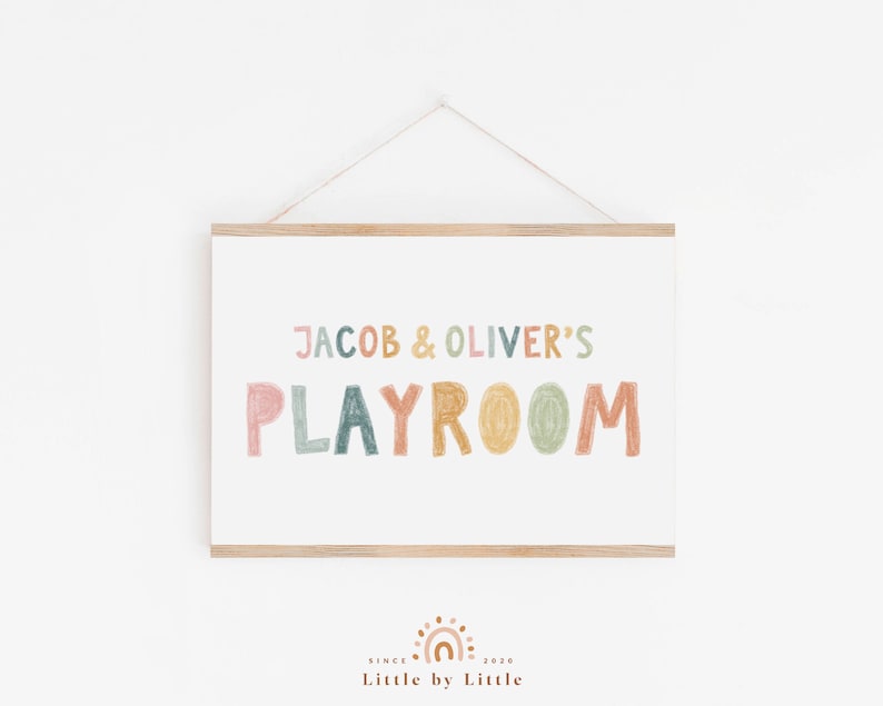 May include: A framed print with the text "Jacob & Oliver's Playroom" in colorful, hand-drawn letters. The print has a light wood frame and is hanging on a white wall.